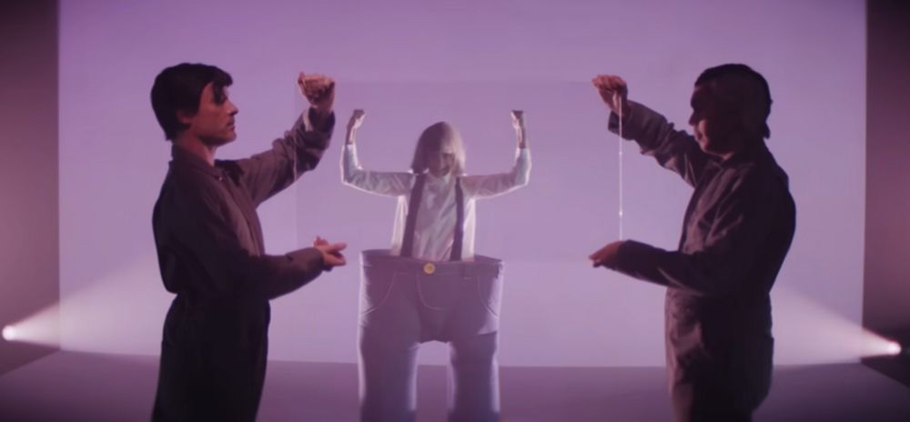 Screenshot from Sia’s music video “Unatoppable” where she’s wearing a black and white wig and goofy oversized trousers, smashing glass windows over and over. 