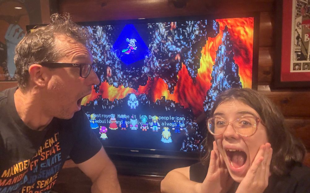 My daughter is in the foreground with a 😱 I’m looking at here with a 😱 In the background is a large TV with the final scenes before the boss fight with Kefka at the end of FF6.