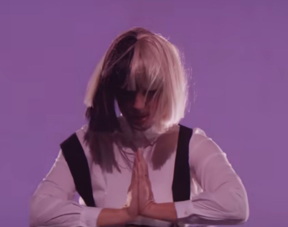 Screenshot from Sia’s music video “Unatoppable” where she’s wearing a black and white wig and goofy oversized trousers, smashing glass windows over and over. 
