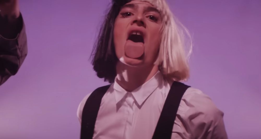 Screenshot from Sia’s music video “Unatoppable” where she’s wearing a black and white wig and goofy oversized trousers, smashing glass windows over and over. 