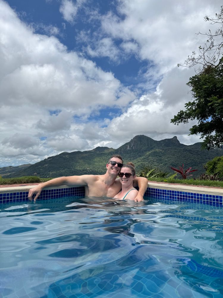 My wife and I are smiling, in sunglasses, and in chest-deep water in corner of a swimming pool and have my arm around her. In the background is the forested mountains of Panama. 