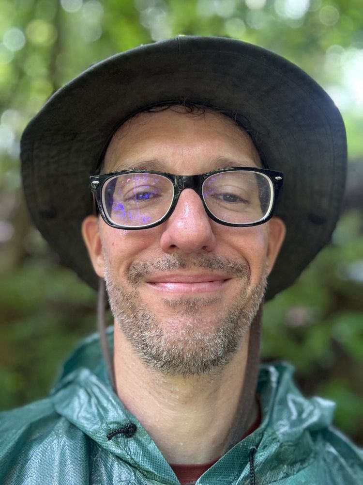 A close up selfie of me in poncho and hat in the rain, in the background is the forest.