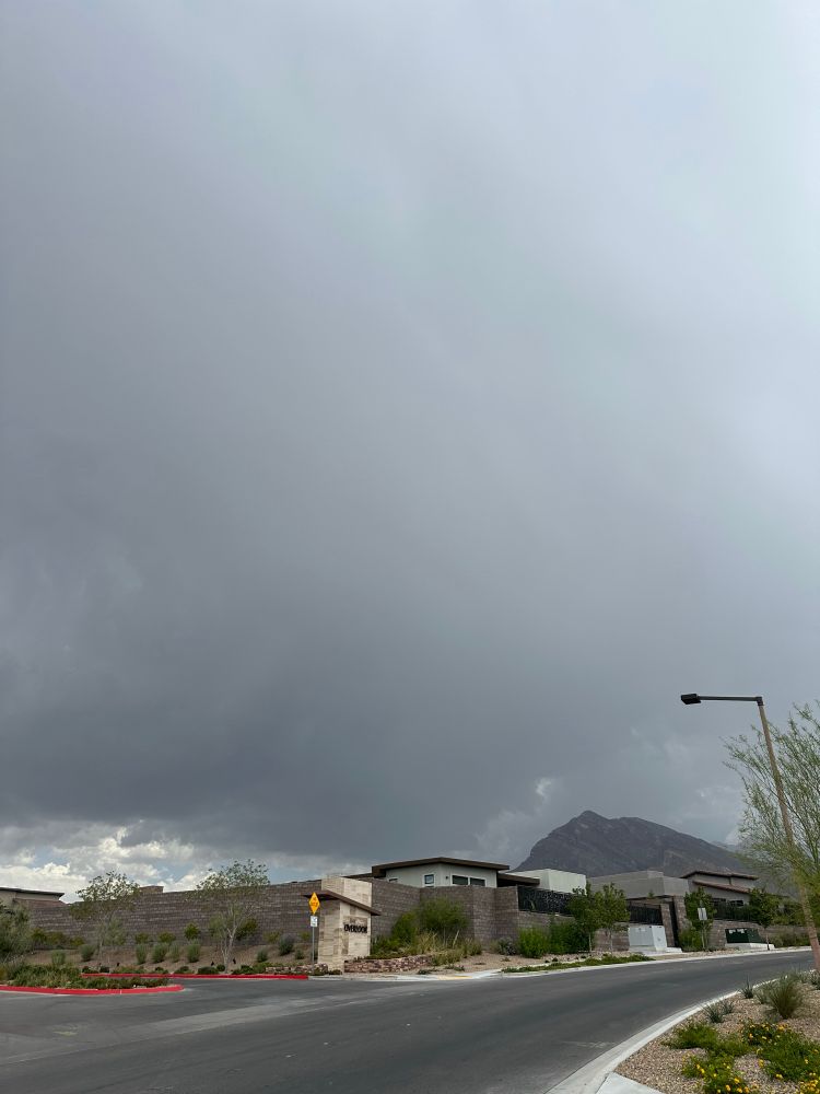 Dark and stormy clouds visible over a desert neighborhood with tall mountains in the background.