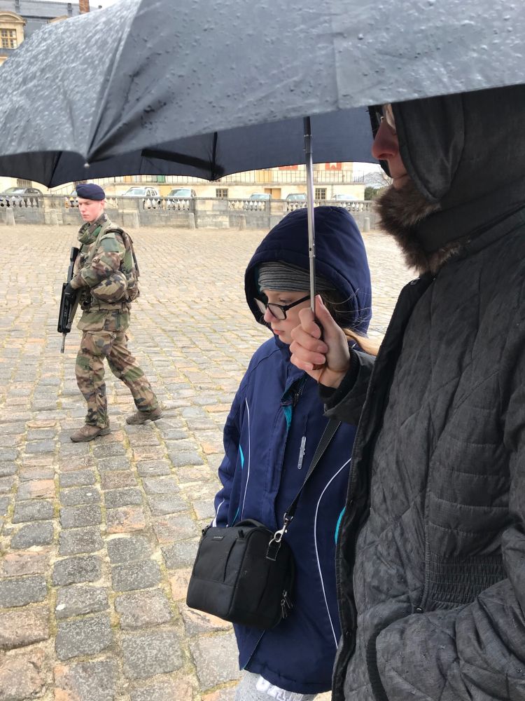 Mother and daughter sharing an umbrella on the cobblestone courtyard of the palace of Versailles. In the background, there is a young soldier carrying a big assault rifle. 