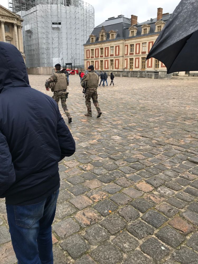 At the cobblestone courtyard of the palace of Versailles. In the background, there is a pair of young soldiers carrying big assault rifles. 