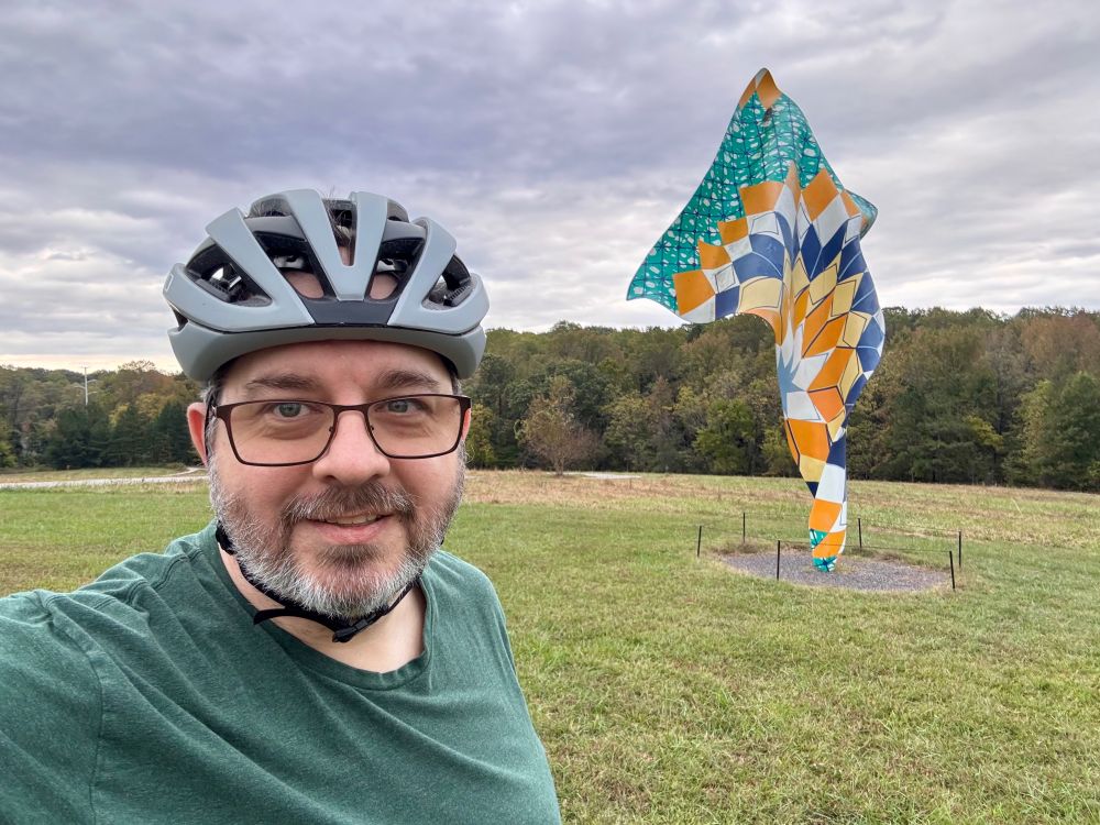 Selfie of Magnus in front of a sculpture installed in a grassy field. Magnus is wearing a bicycle helmet.