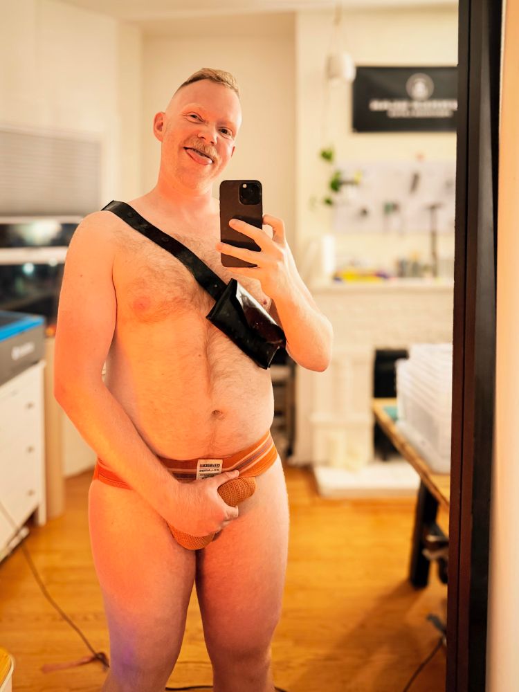 Man standing in front of a mirror wearing nothing but an orange jockstrap and a black rubber cross-body bag from Bunker Gear Precision Latex holds his crotch, the outline of his thick engorged cock visible through the strained mesh of his jockstrap. Behind him, out of focus, is a workroom brightly lit with various tools and the end of a workbench visible. He is lit from the front by an orange glow.