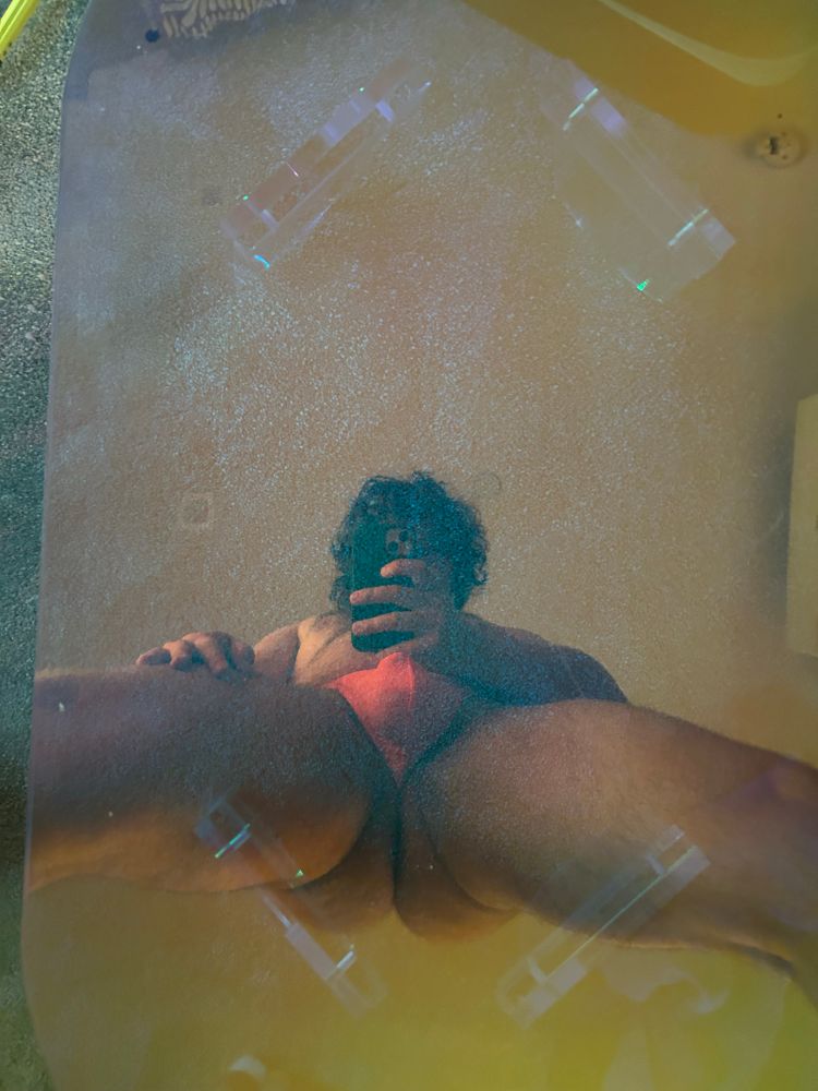 A reflection of a man in an orange thong straddling an iridescent table.  
