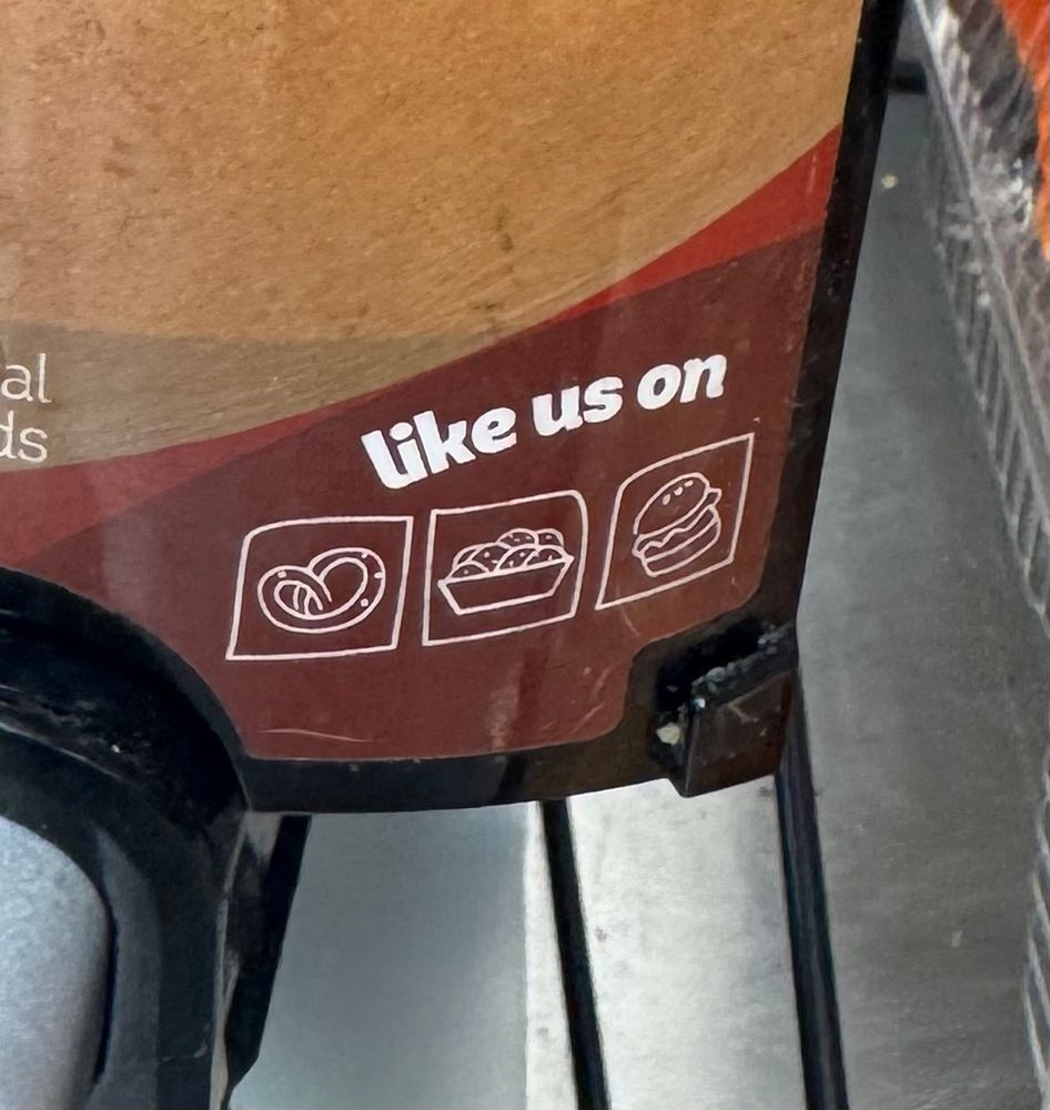 The corner of a chili dispenser saying “like us on” with a logo of a pretzel, nachos, and a hamburger.