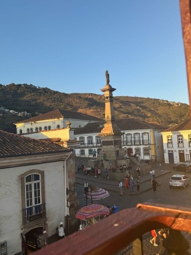 Vista de um sobrado em Ouro Preto-Mg com monumento em homenagem a Tiradentes, na Praça com seu nome, e montanhas da cidade ao fundo com um belo céu azul.
