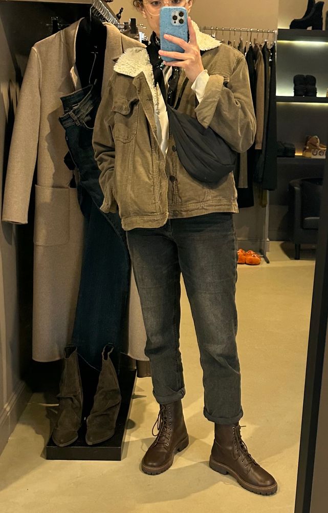 Foto de corpo inteiro no espelho de uma mulher branca, cabelos escuros e ondulados presos num rapinho. Vestida de uma jaqueta de veludo verde oliva com gola de sherpa e por cima uma shoulder bag preta transpassada, uma calça jeans preta estonada e um coturno marrom escuro.