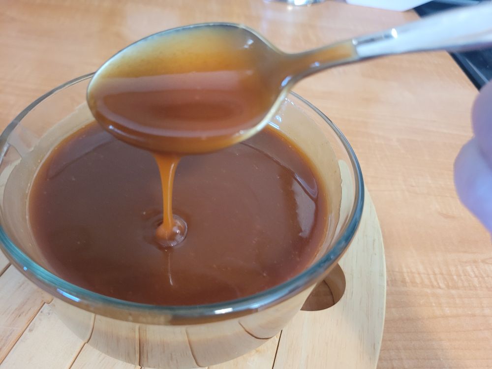Rosemary caramel sauce drips off a teaspoon into a glass bowl. The bowl is sitting on a wooden trivet on a counter. 