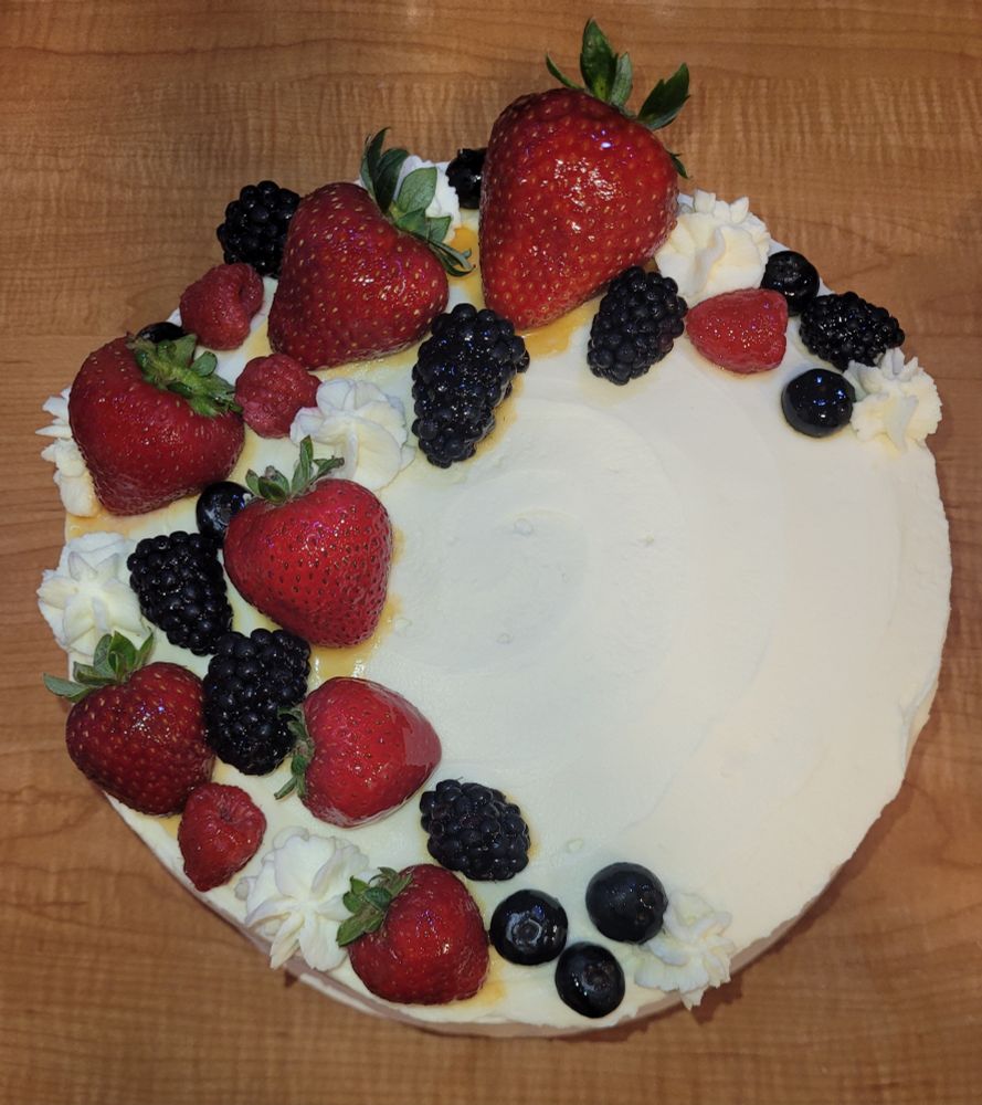 Top view of a white Chantilly cake decorated with a crescent-shaped swath of strawberries, blackberries, blueberries, and raspberries, on a wood grain counter.