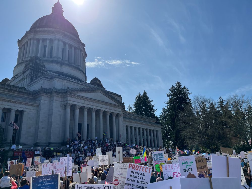 Bad photo of the Olympia, WA Capitol Dome. The stairs are covered in protesters, and protest signs are visible in the foreground with anti-Trump and anti-DOGE messages.
