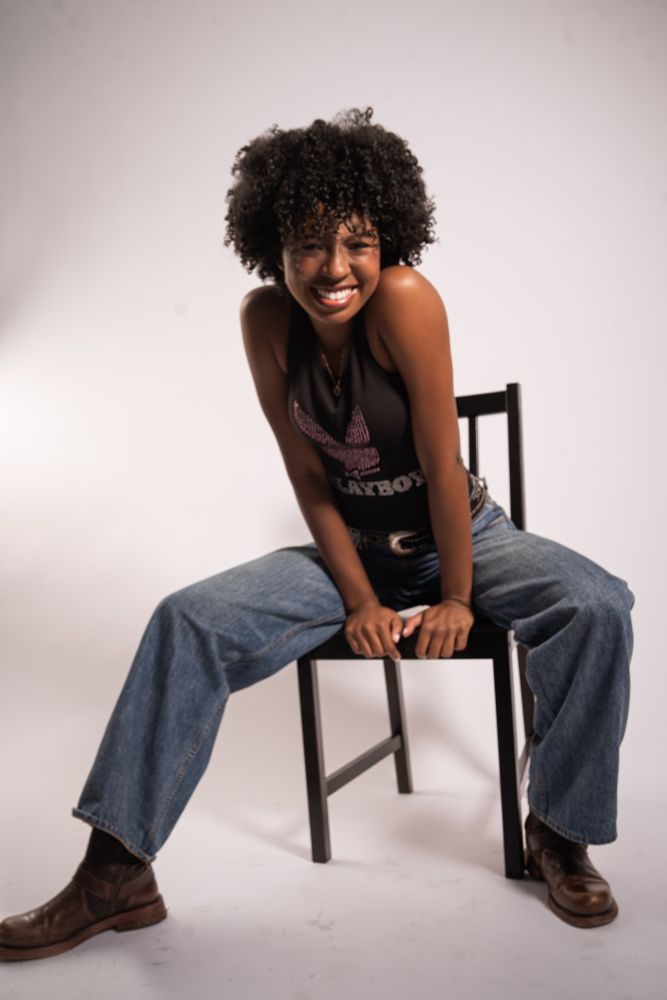 Striking editorial portrait of a poised Black model seated with attitude, styled in wide-leg denim, a fitted tank, and leather boots, framed against a clean studio backdrop.