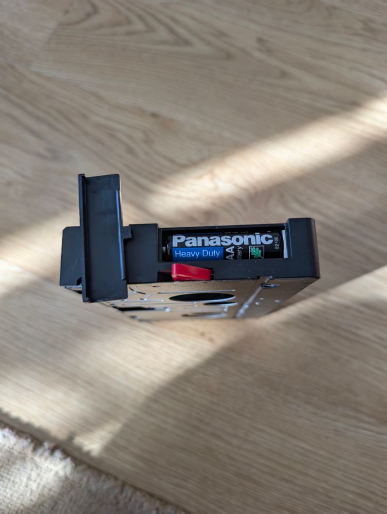 This picture shows the aforementioned Panasonic cassette adapter standing on it's side, with the red piece of plastic protruding in the direction of the camera. The cover for the battery bay is taken off and placed on the side of the cassette adapter itself. Inside the battery bay there's a Panasonic Heavy Duty AA battery with some green leaf logos. There's a fine print on the bottom of the battery: 02±06 *Speculation - it might be the manufacturing date, though the ± sign raises doubts towards that idea