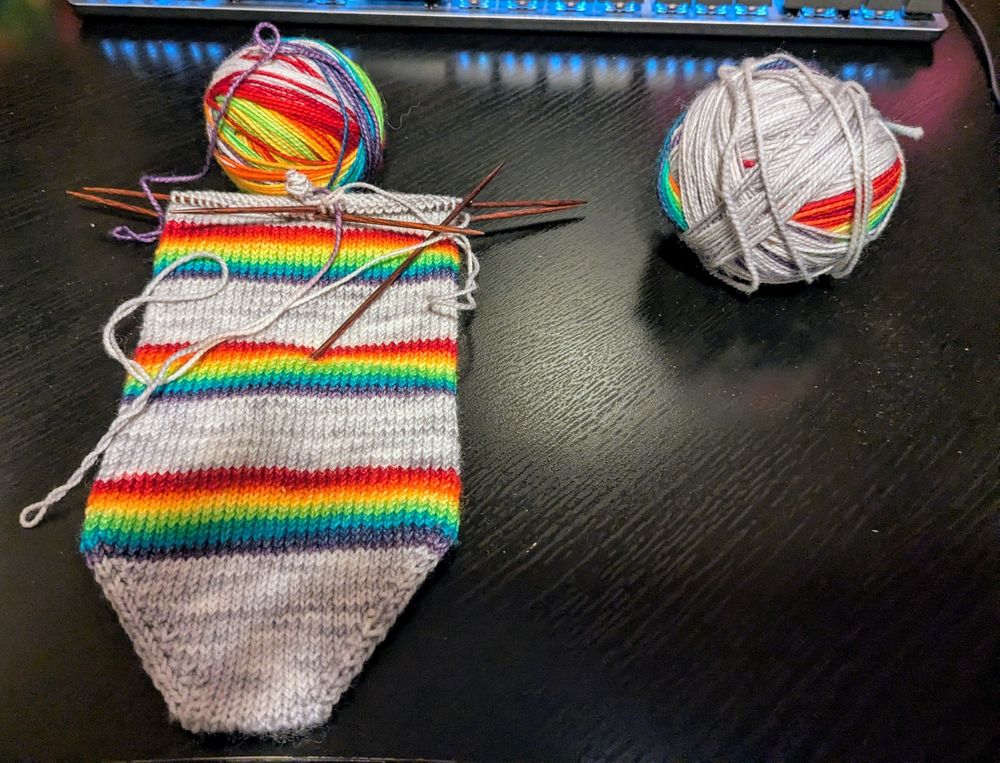 Two balls of yarn, one half complete bottom up sock with alternating gray and rainbow stripes.