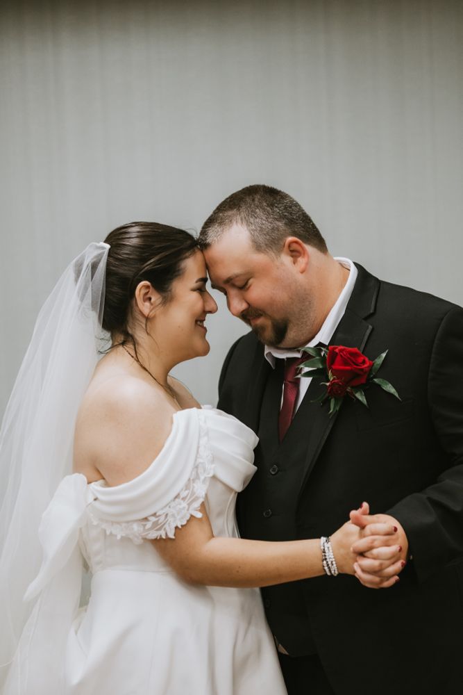 my husband and I on our wedding day - I am in an off-the-shoulder ivory gown and veil, and husband is in a black suit with a red rose boutonniere 
