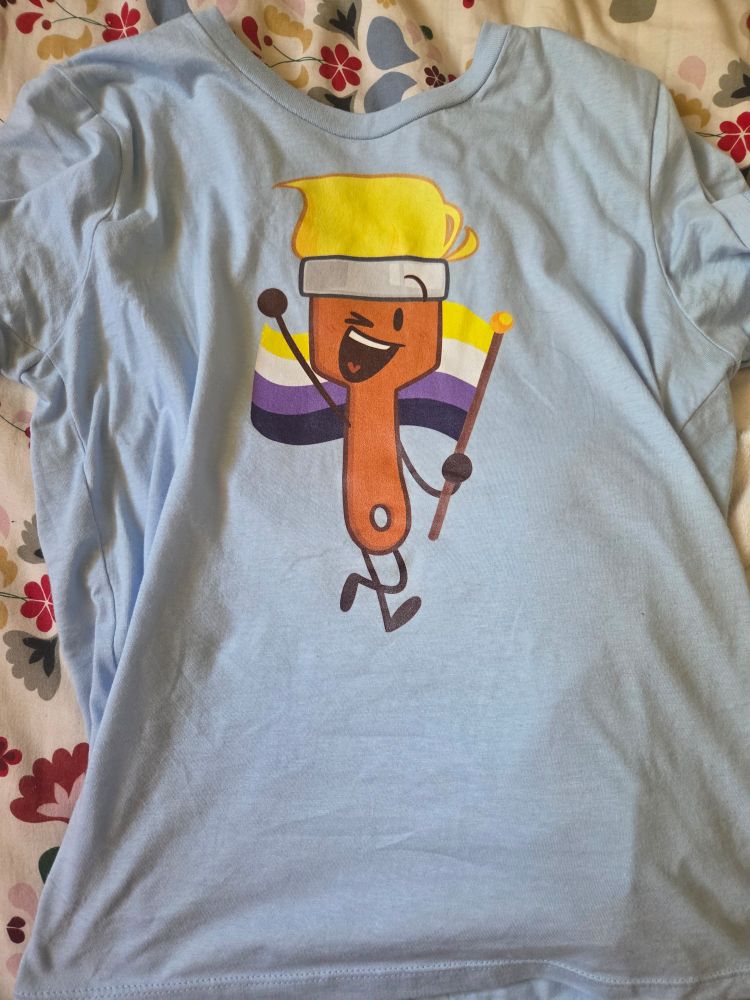 A picture of a t-shirt featuring an image of the character Paintbrush (a brown wooden paintbrush with golden yellow bristles). They are smiling happily with a wink and holding the non-binary flag (horizontal stripes of yellow, white, purple and black).