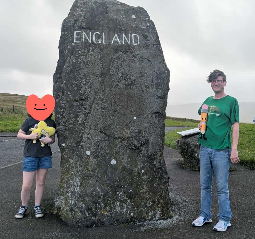 A picture showing me and my kiddo stood either side of a rock with the word "England" etched into it (a spot on the border between Scotland and England). My kiddo is stood on the left of the rock holding a plushie of the character X from Battle for Dream Island (a yellow X) while I am stood on the right side of the rock holding a plush of Pencil from Battle for Dream Island (a yellow pencil with a pink eraser for the top of her head).