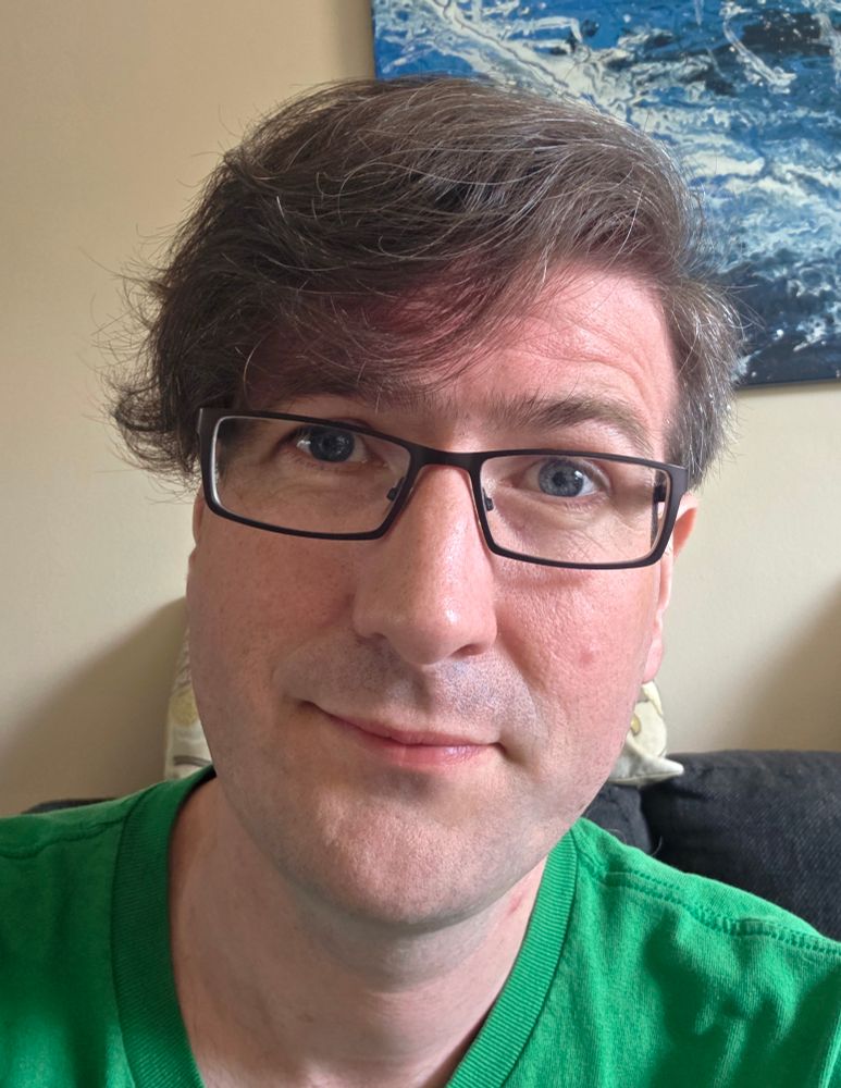 A selfie of me, wearing my glasses and a green shirt. My hair is parted at the side, short on the side and longer on the top.