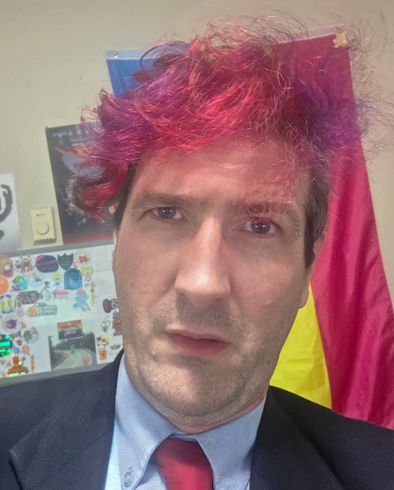A selfie of me looking rather tired and bedazzled. I'm wearing a black suit jacket, blue shirt and a red tie and my hair (which is brown with purple and red in the front) is quite a mess.