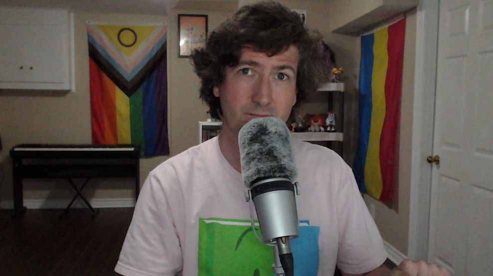A picture of me in my recording room. I'm wearing a pink shirt featuring the character Book from Battle for Dream Island (a book with a half green / half blue cover) and my microphone is in front of me. There is an electric piano in the background to the left of me and the progress pride flag is hung above that. The pansexual pride flag is to my right.