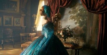 Elizabeth is wearing a feathery blue gown. Her hair is in a bun and she has a feather veil blocking the side of her face. She is turned away from the audience holding up a skull and examining it. The background behind her is a beautiful painting of trees with red curtains draped around it. There is a table in front of the painting with a case of flowers and some books. 