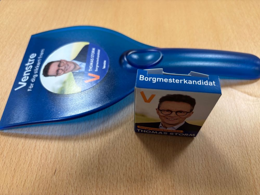 Blå isskraber og lille blå papæske i gajolstørrelse på et bord. Begge dele har politisk kampagnedesign for Venstre med portræt af en kandidat (Thomas Storm), partifarve og tekst om borgmesterkandidatur.