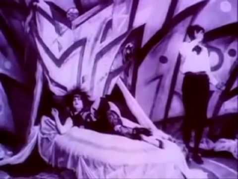 Purple film still from Genuine (1920) with a woman on a bed and a man standing with his right hand over his face 