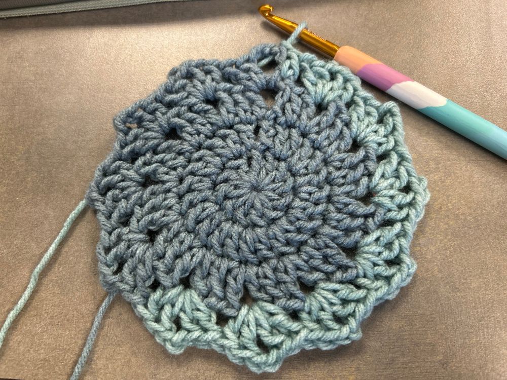 Beginnings of a crochet blanket using an ombre acrylic yarn in a grayish-green color. Golden crochet hook with polymer clay handle sits in the top right corner, hooked onto the working loop of the project.