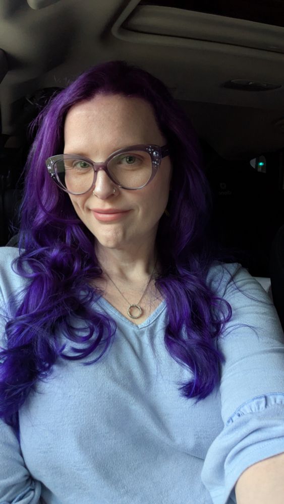 Amy smiles wearing a blue shirt with her purple hair down and wavy.
