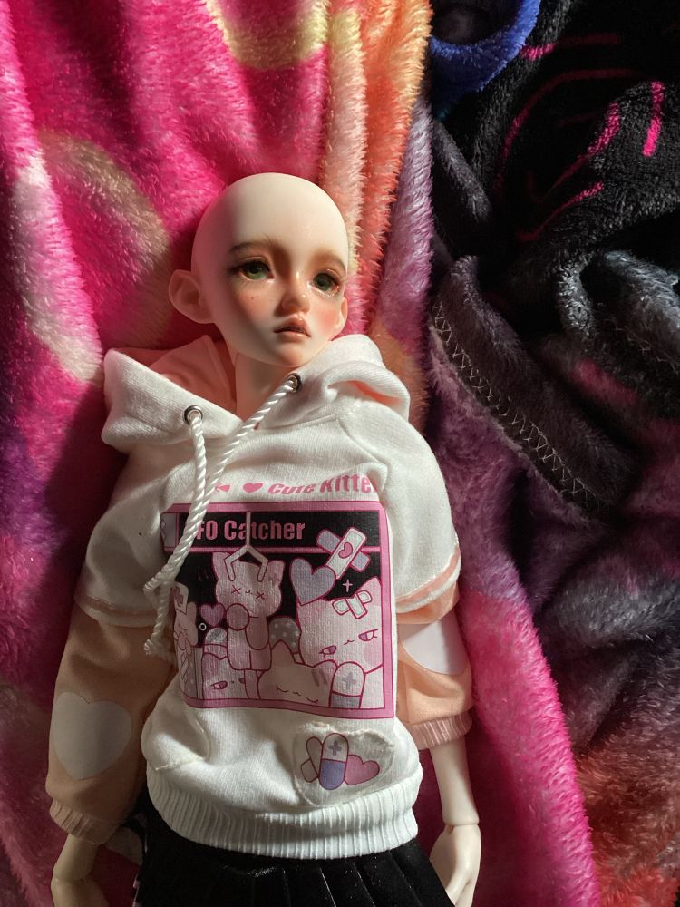 A photo of my bjd Alaric. He is wearing a pink cat ear hoodie with a black skirt. He is bald and has grey green eyes. (These things will change with time.) 