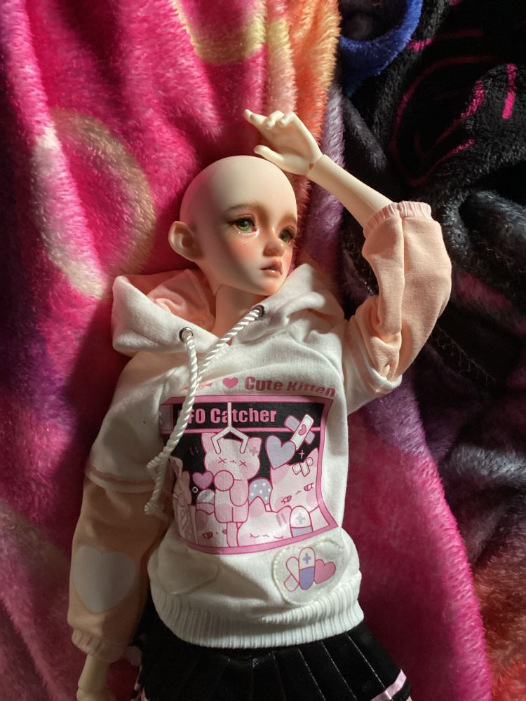 A photo of my bjd Alaric. He is wearing a pink cat ear hoodie with a black skirt. He is bald and has grey green eyes. (These things will change with time.) 