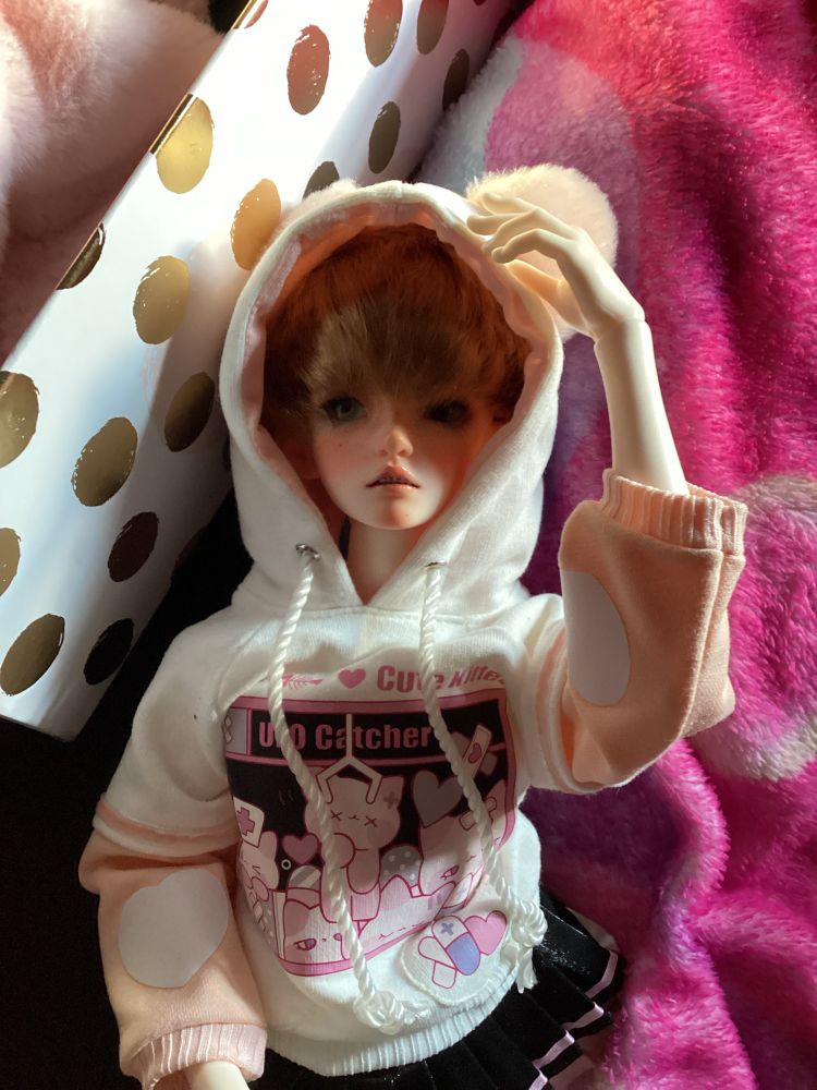 A photo of my bjd Alaric. He is wearing a pink cat ear hoodie with a black skirt. He has short dark brown hair and grey green eyes. (These things will change with time.) 