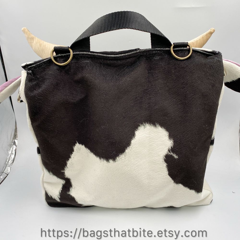 The back of the soft cow briefcase showing off the soft faux fur in black and white cow patch, as well as a better view of the top fabric handle and the top sewn in D rings to connect a shoulder strap. Side d rings also are sewn in if the adjustable removable strap is preferred to be attached on the sides. 