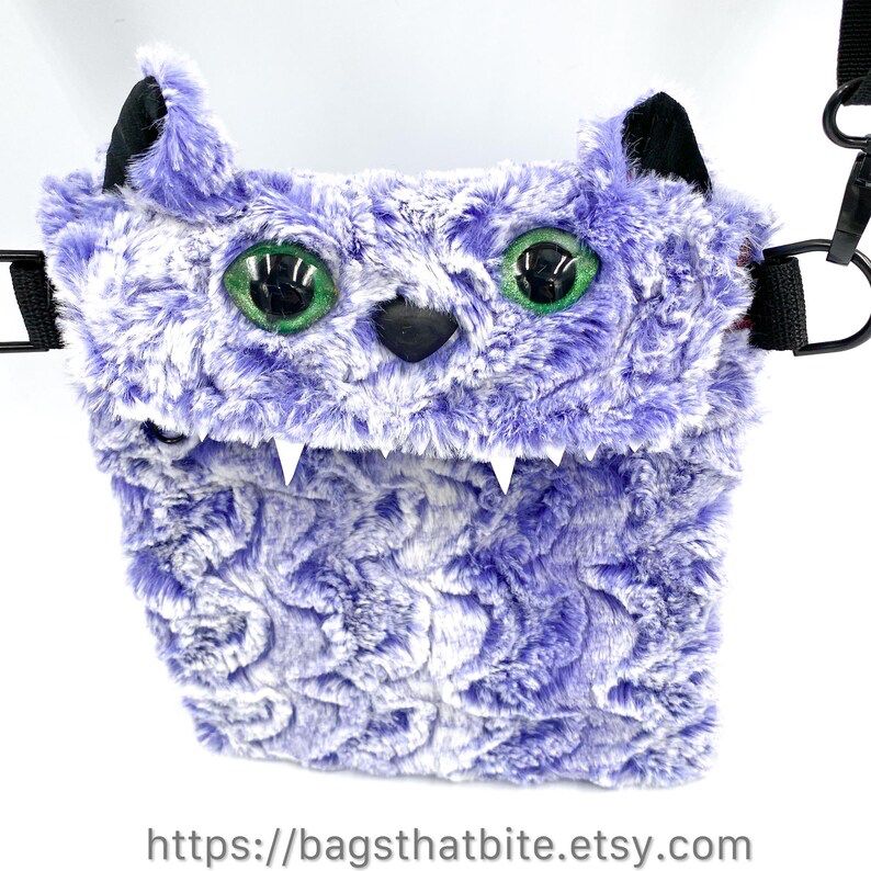 Purple and white wavy fur (that is very soft!) rectangle monster bag with cat ears, green cat eyes and soft black triangle nose. D-Rings on side allow included adjustable shoulder strap to carry bag. 