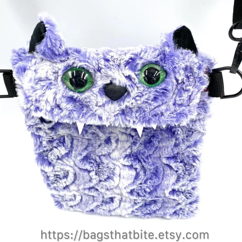 Crossbody bag in lilac faux fur with almond shaped green cats eyes, cat ears, cat nose and pointy foam teeth. 