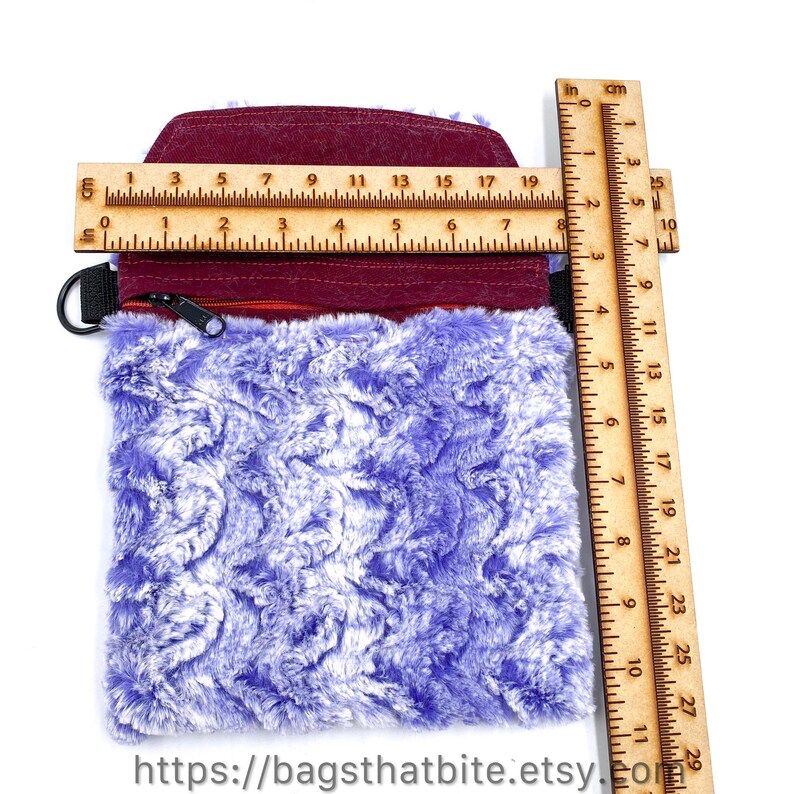 Measurements of interior of zipper bag are 7 3/4” across and 7 1/2” deep. 
Purple and white wavy fur (that is very soft!) rectangle monster bag with cat ears, green cat eyes and soft black triangle nose. D-Rings on side allow included adjustable shoulder strap to carry bag. 