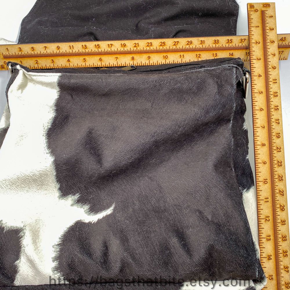 A large rectangular bag  in a cow faux fur is open with rulers to show the size of the bag. The zipper opening along the top is 13” across and the height of the bag is 12”. 