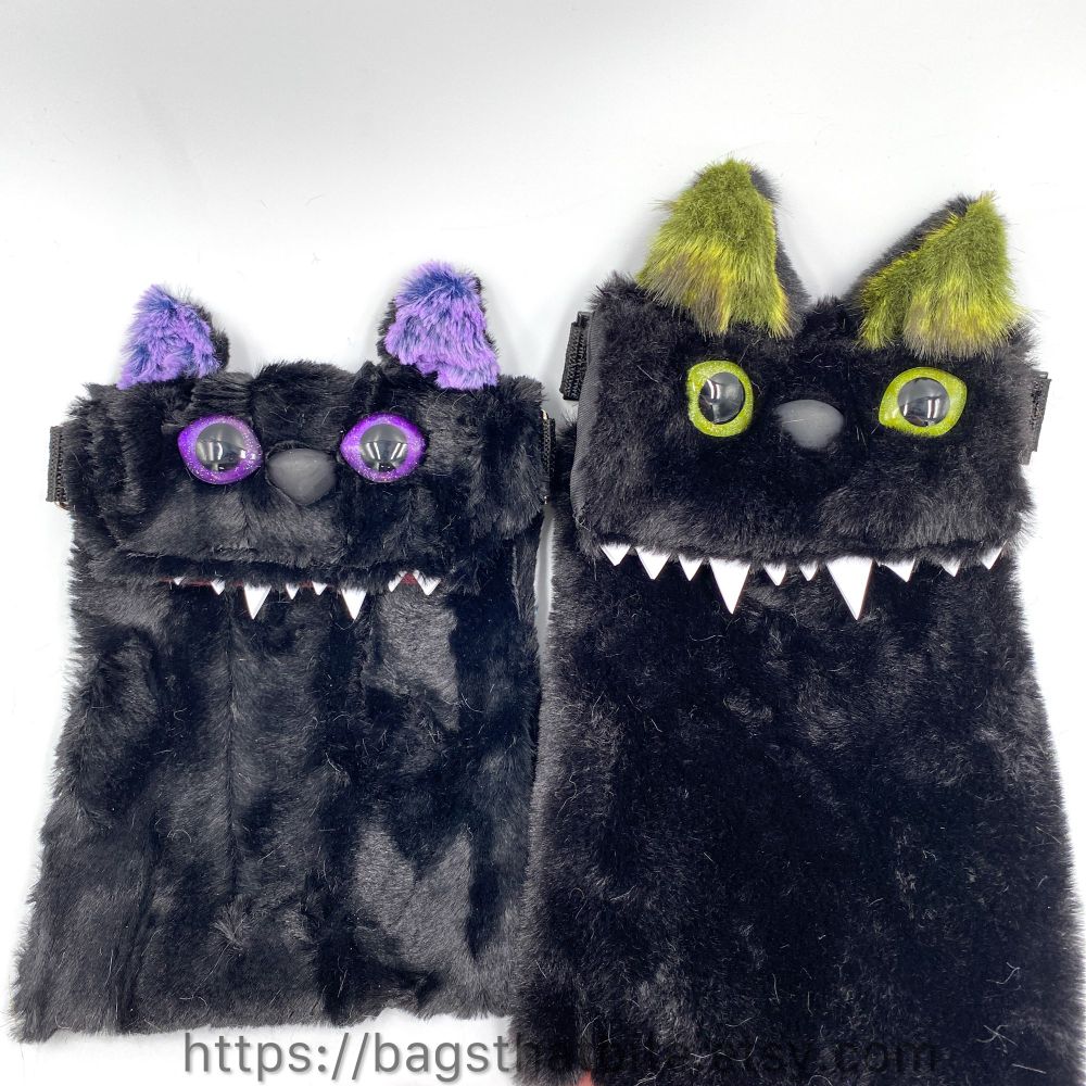 2 black faux fur rectangle crossbody bags with cat ears, hand painted eyes and pointy foam teeth. The bag on the left has purple eyes and purple inner ear. The bag on the right has green eyes and green inner ear. 