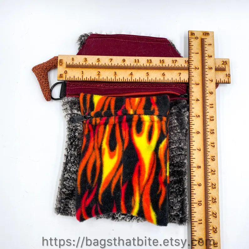 This is a crossbody (or shoulder) monster bag that is made from a faux fur of marbled gray and white with crocodile scales as a pattern. It looks fierce, but is butter soft to touch. This is a photo of the mouth flap flipped up  with rulers next to the zipper closure to show the dimensions of the bag. (7” wide x 11” tall; the storage portion of the bag being 8” tall). It also shows the attached forked flame tongue that is a zipper pocket that can hang out of the unzipped mouth or tucked in to zip the bag closed. 
