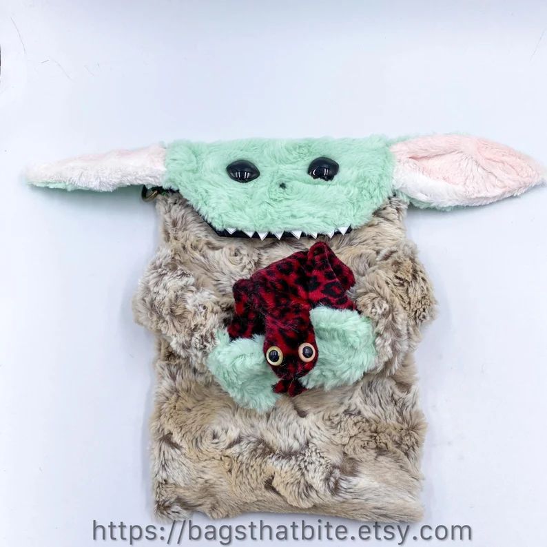 A bag that resembles a baby space goblin that may or may not be from a popular sci-fi show. He is holding with his bendable fingers, a zippered change purse, red leopard alien frog.
