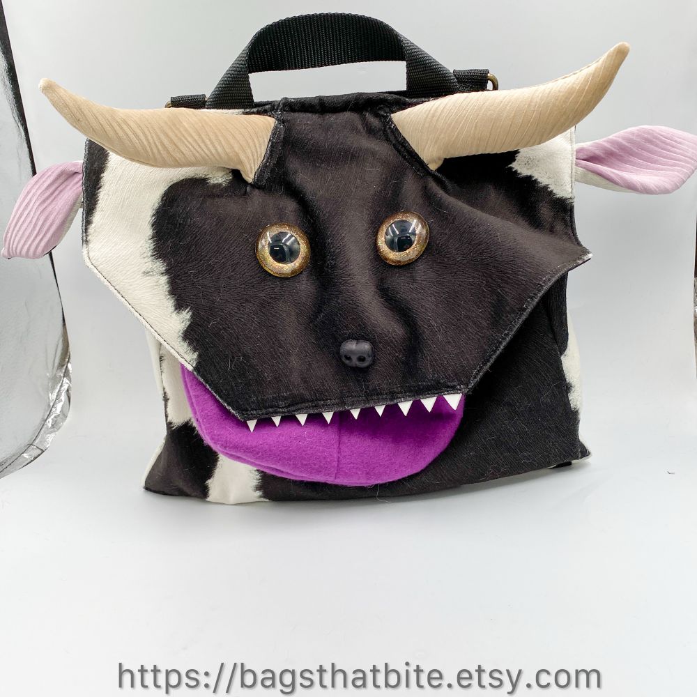 A large rectangular bag  in a cow faux fur with large hand painted golden brown eyes, soft nose and pointed foam teeth. A zipper pocket tongue bleps. Large fabric horns and pink lined ears and fabric handle finish the look. 