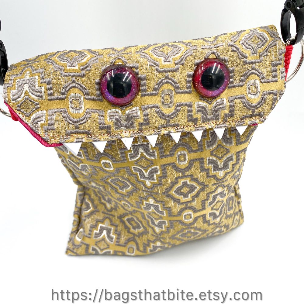 Tan rectangular monster bag with hand painted red purple sparkling eyes, white foam teeth and two tone silver geometric embellishments embroidered on the fabric. 