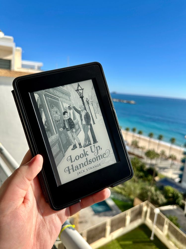 Look Up, Handsome by Jack Strange is shown on a Kindle e-reader. The photo is taken from a balcony and in the background there is a beach, ocean, and palm trees. 