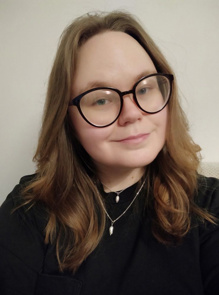Selfie of a white, blonde person. They are wearing a black t-shirt and two pearl necklaces. They have large black glasses and are smiling a little at the camera.