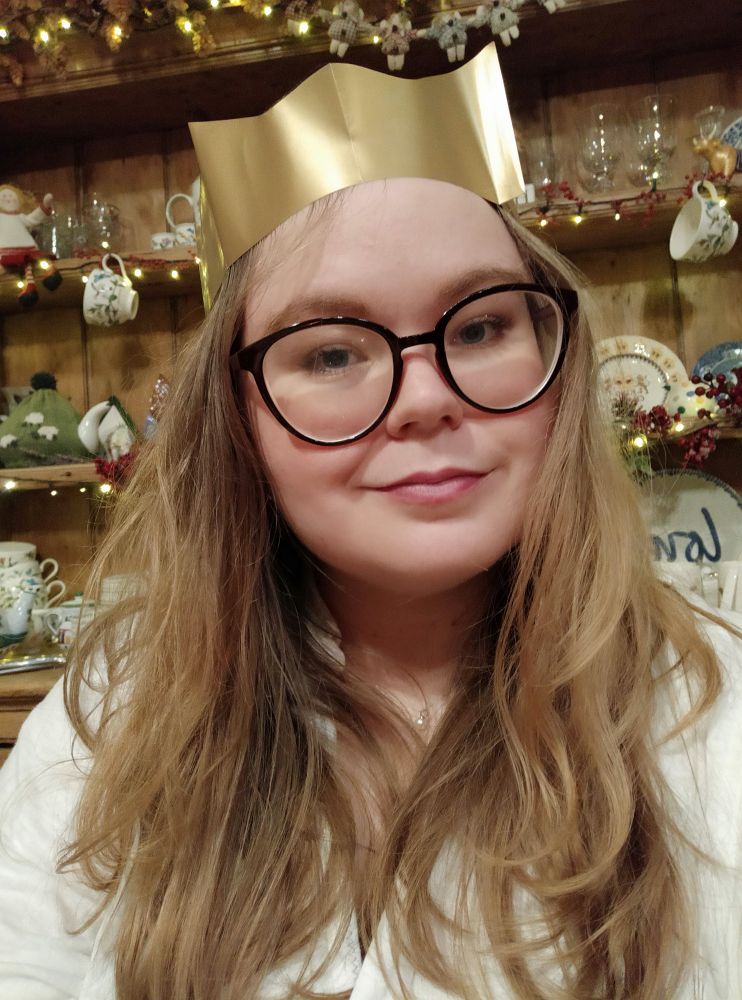Selfie of a white person with long blonde hair. They are wearing a gold paper crown from a Christmas cracker. Their expression is happy but a little drunk.