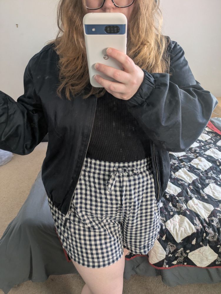 Mirror selfie of a white person with long blonde hair. They're holding a white phone in front of their face and wearing short black and white gingham shorts and a black bomber jacket.