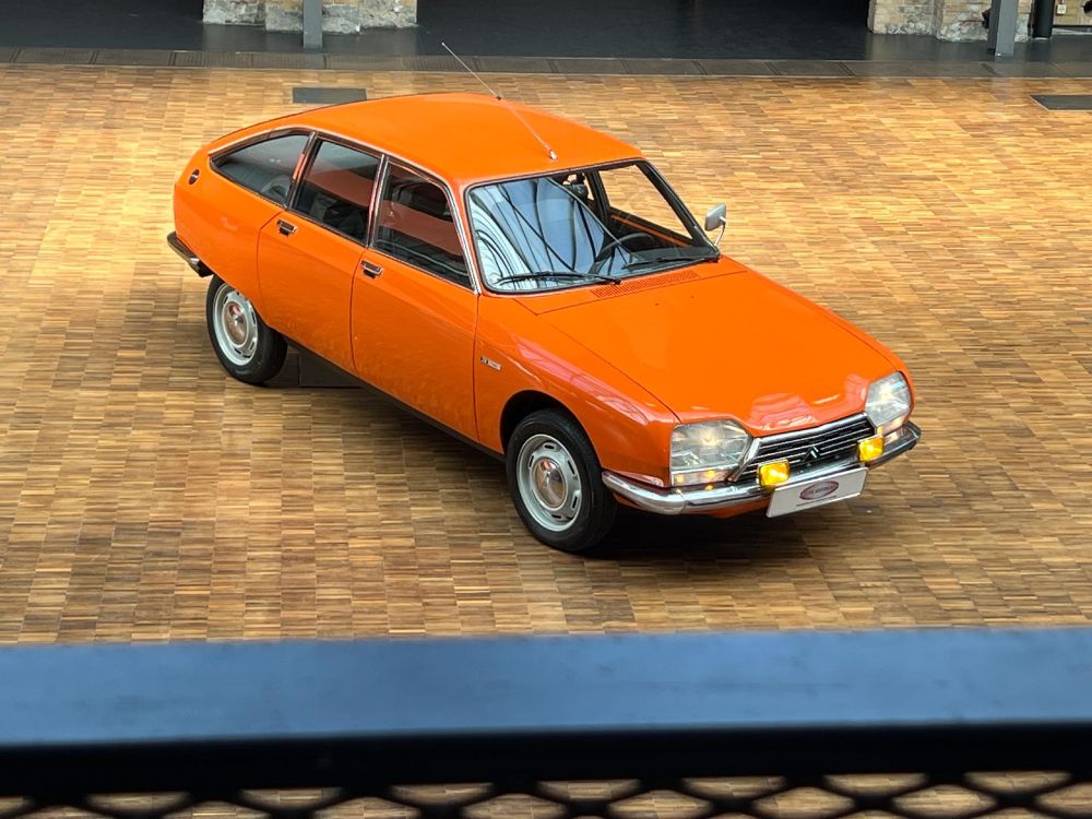 Der orangefarbene Citroën von halb vorne und oben: große fünfeckige Scheinwerfer und gelbe Nebelleuchten, praktische Fließheckform mit vier Türen, die auch hinten einen großen Ausschnitt aufweisen, verkleideter hinterer Radkasten, einfache Felgen mit Chromnabenkappen und schmalen Reifen
